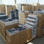 Pilhas de mercadorias irregulares apreendidas em armazém no DF, com arquitetura de Brasília ao fundo.