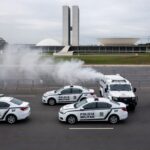 Cena de confronto com viaturas da PM no DF, fumaça de spray de pimenta no ar e monumentos de Brasília ao fundo.