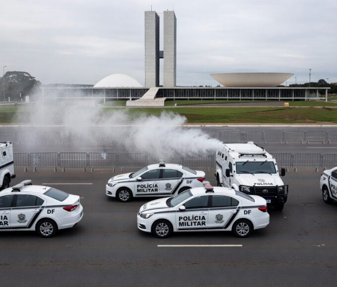 Cena de confronto com viaturas da PM no DF, fumaça de spray de pimenta no ar e monumentos de Brasília ao fundo.