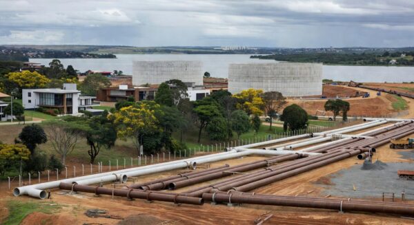 Obras de ampliação de água no Lago Sul, Brasília, com tubulações e Lago Paranoá ao fundo, em meio a críticas por atrasos.