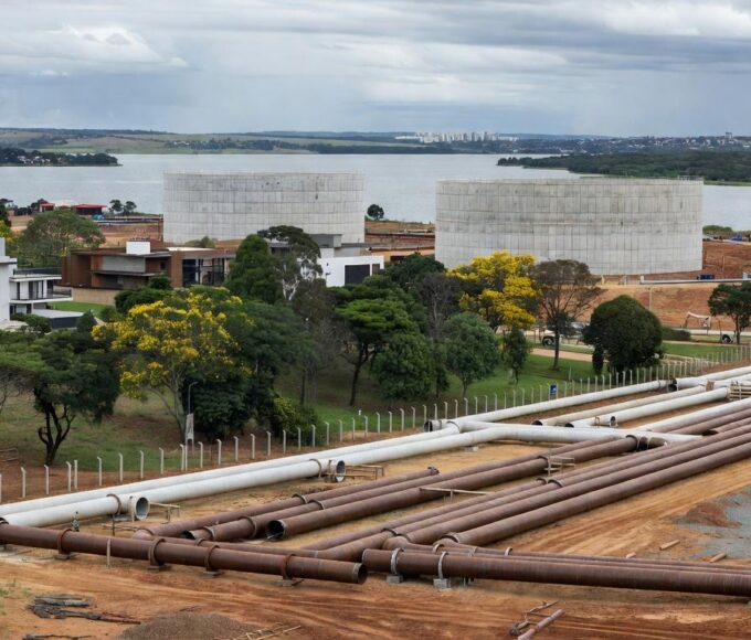 Obras de ampliação de água no Lago Sul, Brasília, com tubulações e Lago Paranoá ao fundo, em meio a críticas por atrasos.