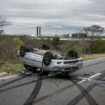 Carro capotado em acidente na DF-005 em Brasília, com destroços na rodovia e vegetação do cerrado ao fundo.