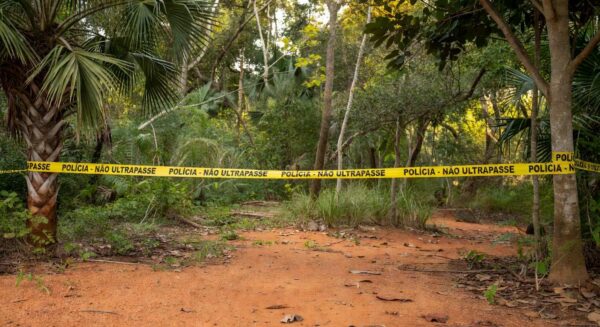 Cena de crime isolada com fita policial em floresta brasileira no Distrito Federal, representando denúncia por latrocínio e ocultação de cadáver.